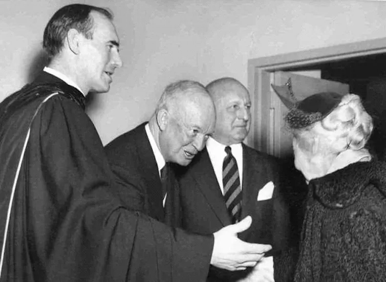 Three men, two wearing black suits and one wearing a priest's robe, greet a woman.