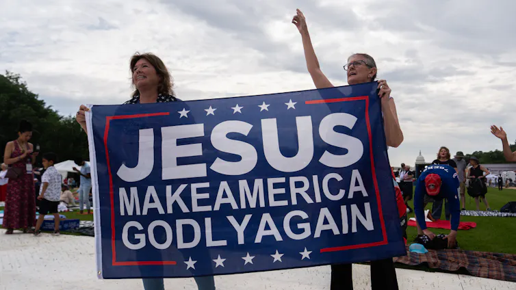Two people hold a large sign that reads, 'Jesus Make America Godly Again.'