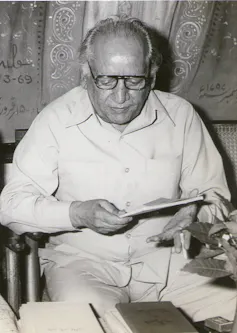 Black-and-white photo of a balding, portly man who's seated, wearing glasses, and glancing down at a notebook.