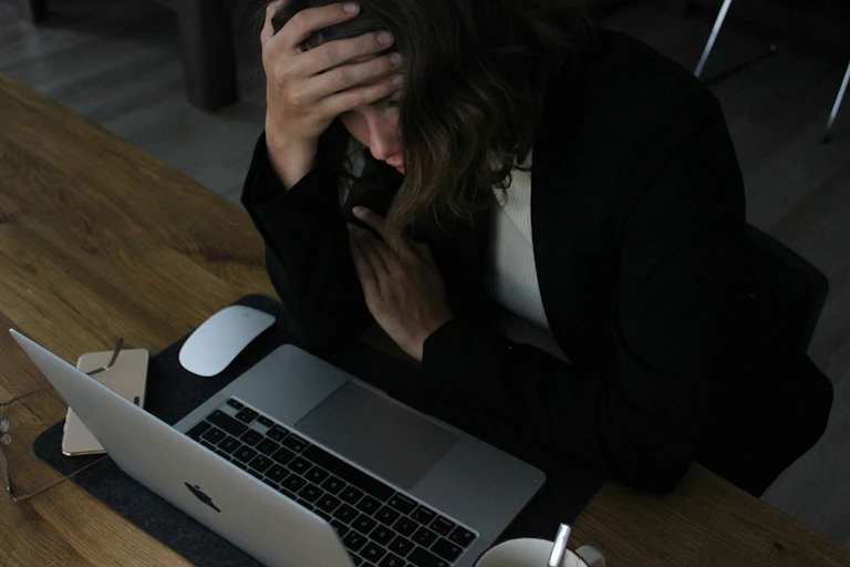 A stressed woman hunches over her laptop, with her right hand on her forehead.