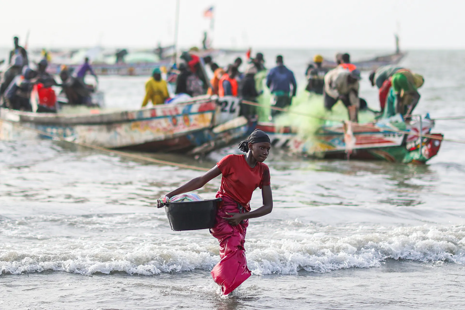 A woman carrying a basket walks out of the surf, with boats behind her.