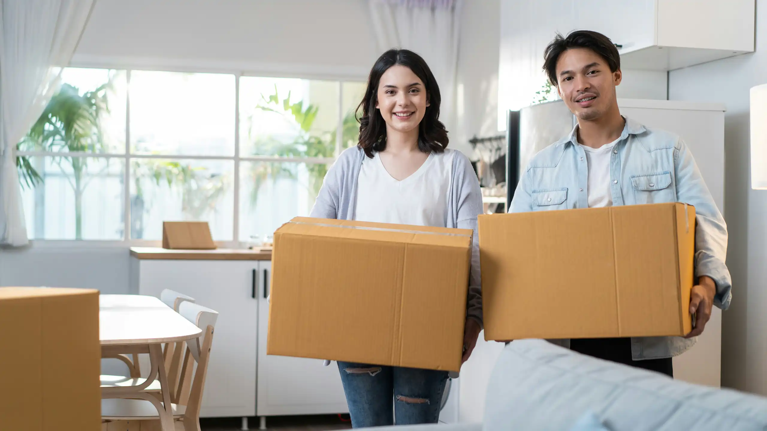 A man and a woman are standing in a the room of a house while holding moving boxes.