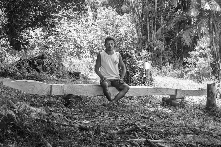 Foto em preto e branco mostra um homem sentado num banco, no meio do quintal