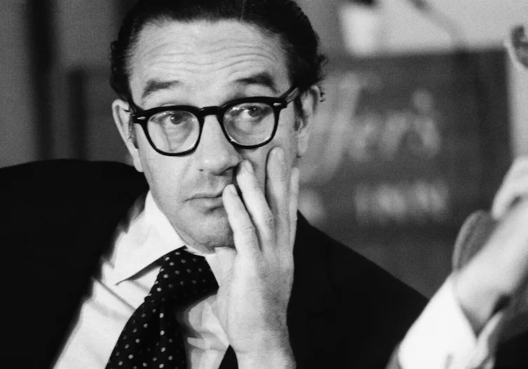 a bespectacled man wearing a suit holds his left hand to his face