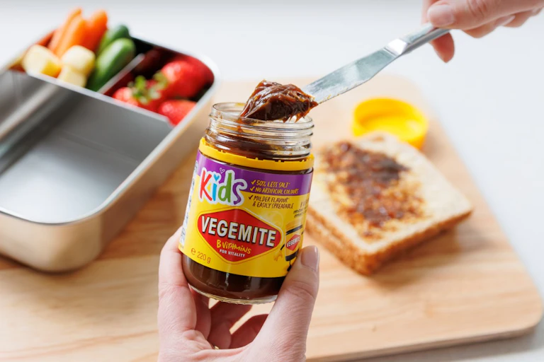 Someone holding jar of Vegemite Kids in the hand, knife with spread on it in other, against background of lunchbox, Vegemite sandwich