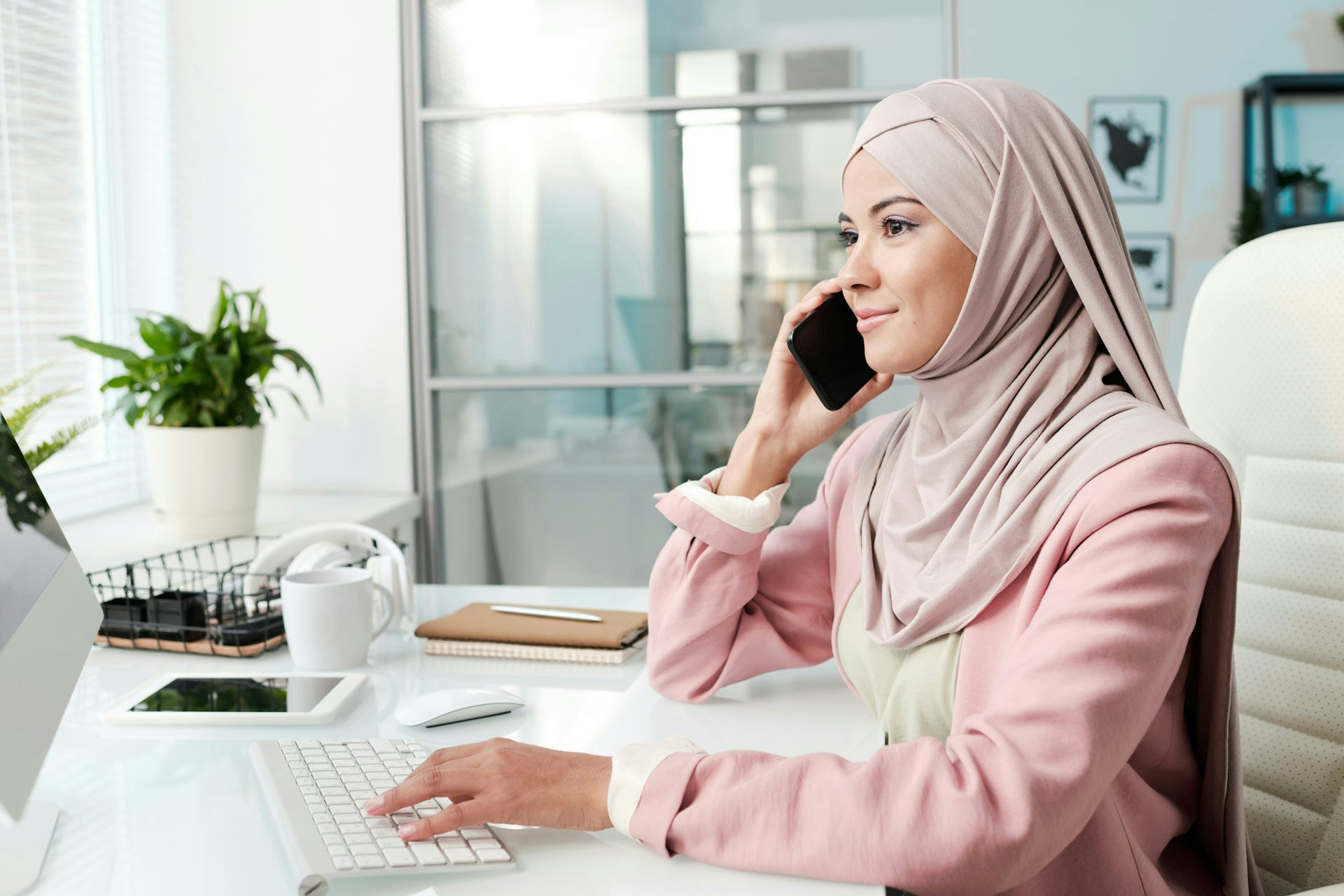 Una mujer sentada en su escritorio en el trabajo contesta el teléfono y escribe en el teclado de la computadora.