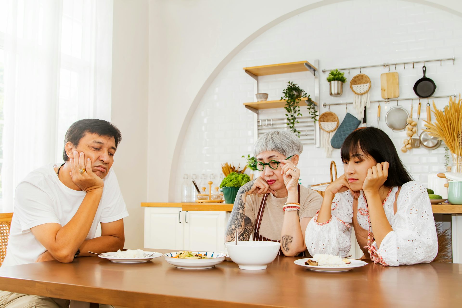 Two older adults and their adult daughter sit around a table of food, looking disappointed or like they've lost their appetite.