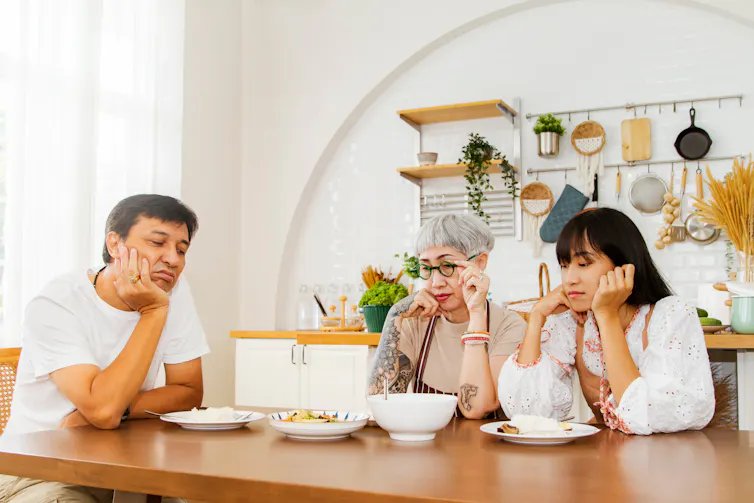 Two older adults and their adult daughter sit around a table of food, looking disappointed or like they've lost their appetite.