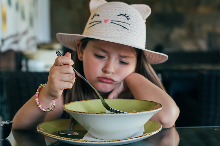 A young girl wearing a cat sun hat sits and frowns at the bowl of food in front of her.