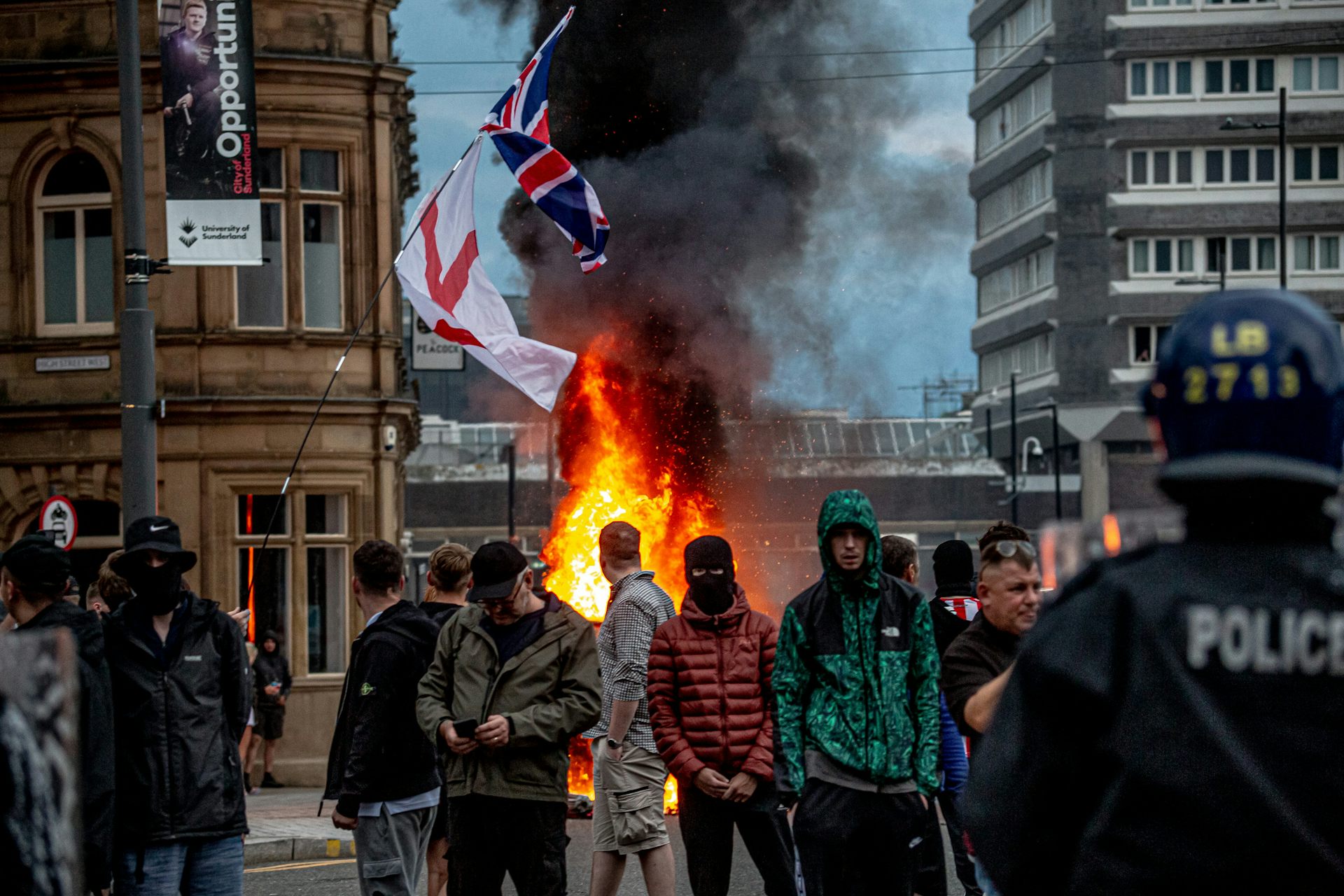 Masked demonstrators stand in front of flags and flames.