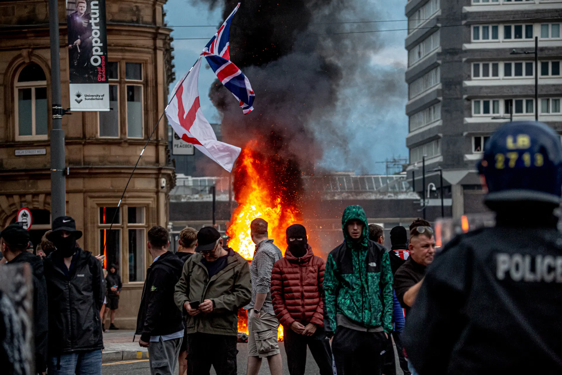 Masked demonstrators stand in front of flags and flames.