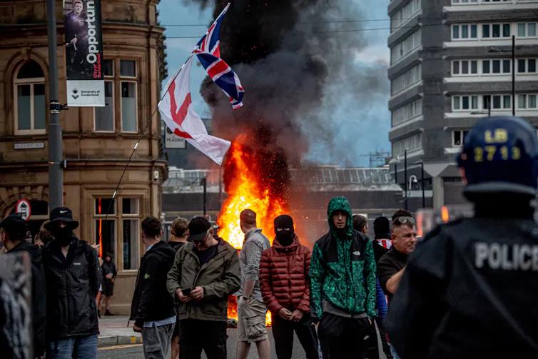 Masked demonstrators stand in front of flags and flames.