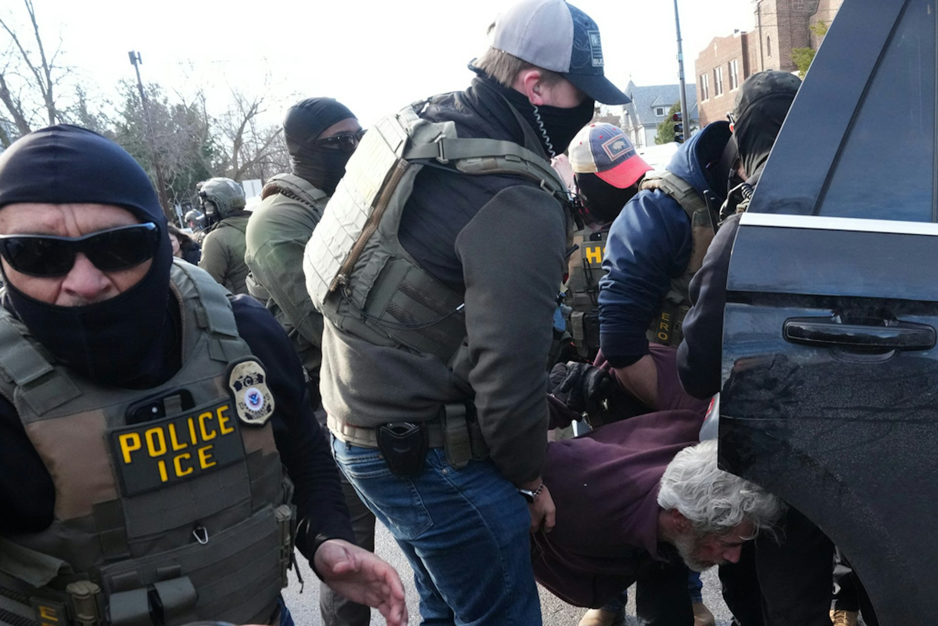 Masked law enforcement agents put someone into the back of a car.