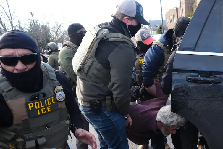 Masked law enforcement agents put someone into the back of a car.