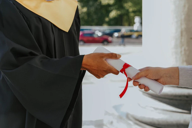 Hands giving and receiving a rolled-up paper bound with red ribbon; one person is wearing an academic gown