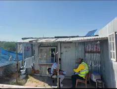 Two people sit outside a corrugated metal shack where a solar panel has been mounted on the roof