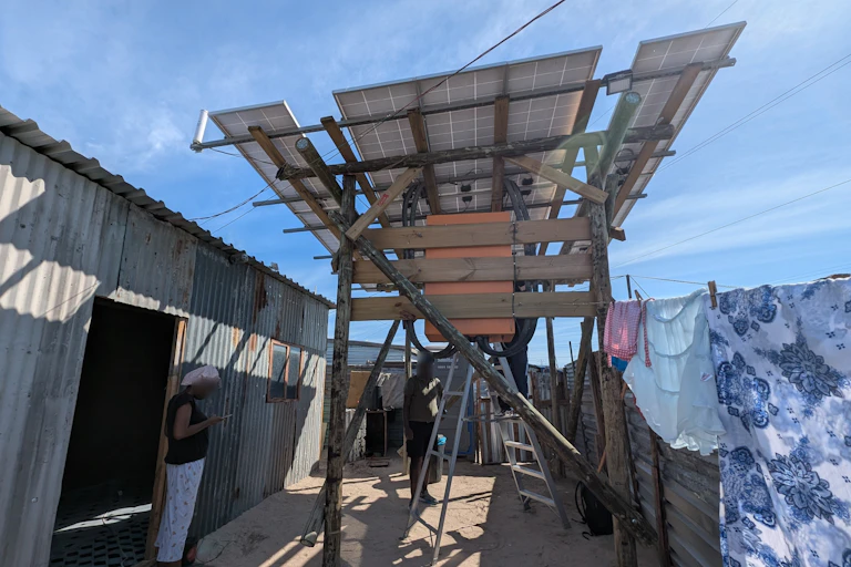 A shack made of corrugated metal with three solar panels mounted on poles outside, generating energy for the residents.