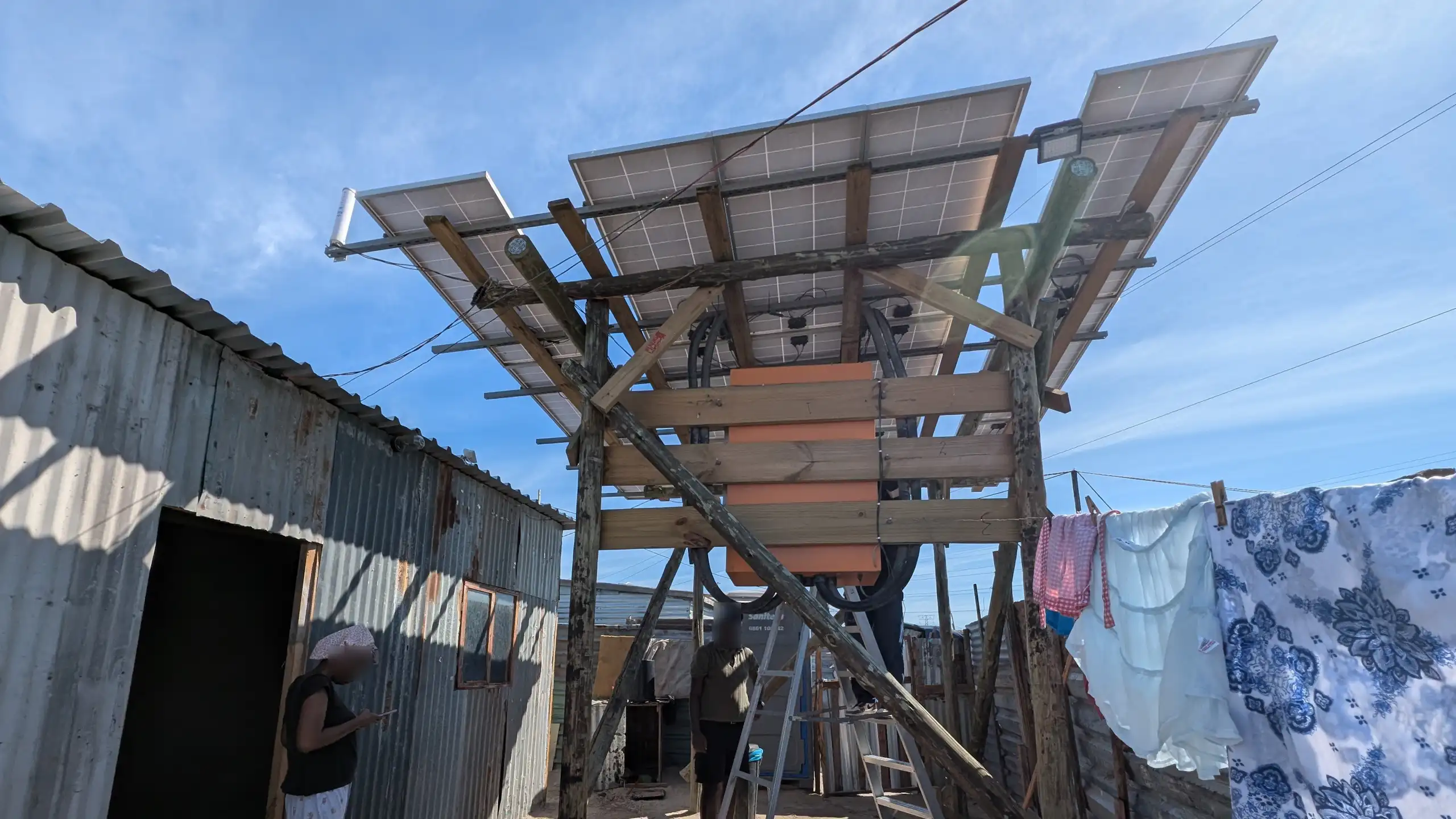 A shack made of corrugated metal with three solar panels mounted on poles outside, generating energy for the residents.