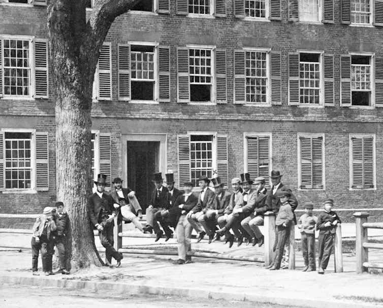 Men dressed formally with top hats stand outside of a large building and next to a tree in a black and white photo.