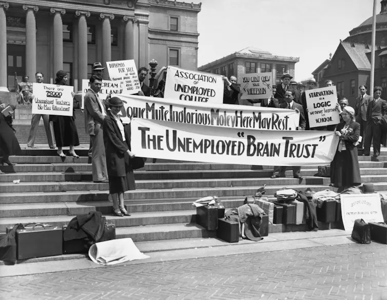 People dressed formally hold signs, including a banner that says 'The unemployed brain trust,' as they stand on steps outside a building with tall columns in a black and white photo.