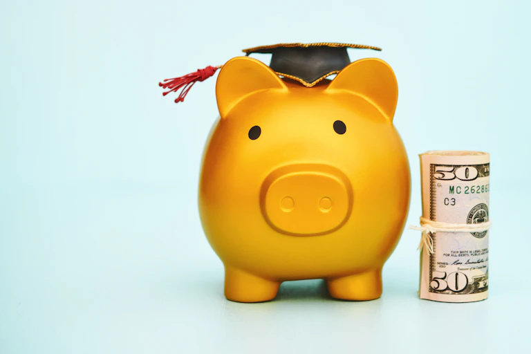 A yellow piggy bank has a rolled-up $50 bill next to it and a black graduation cap on its head.