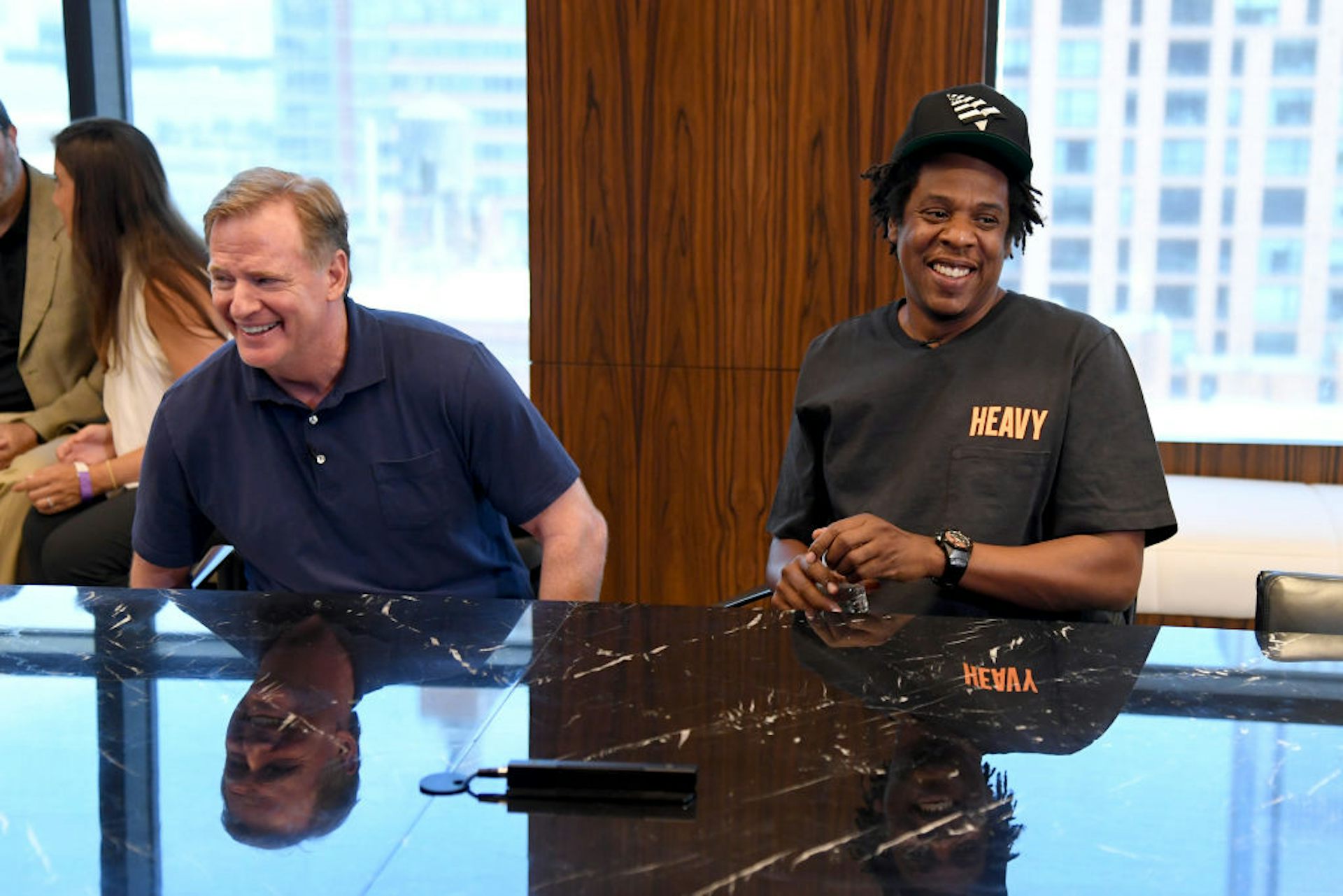 An older blonde man wearing a polo shirt and a Black man wearing a black baseball cap laugh while sitting next to one another.