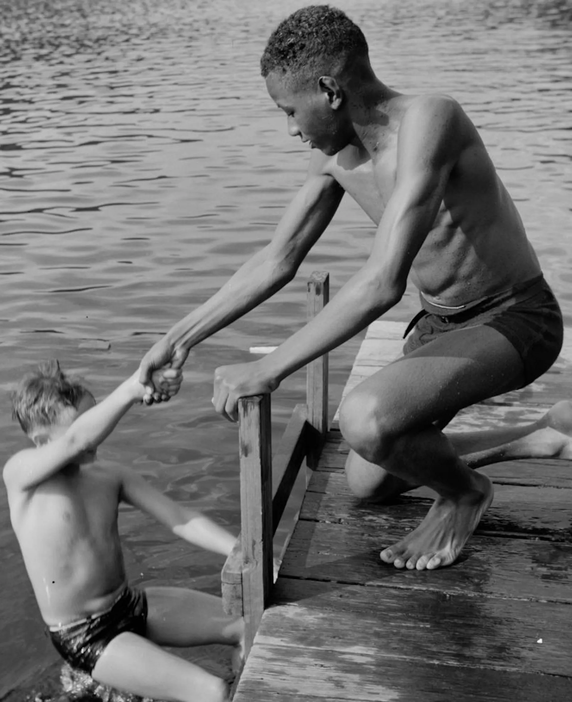 A Black boy stands on a dock and helps lift a white boy out of the water.