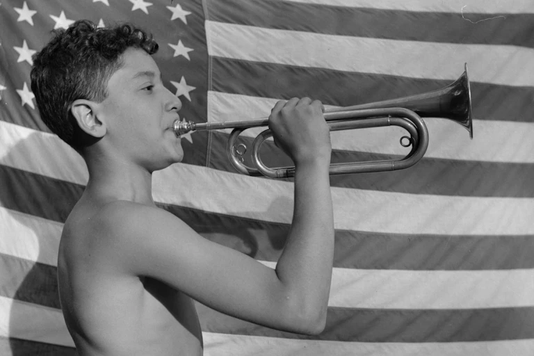 Shirtless boy stands before an American flag while blowing a bugle.