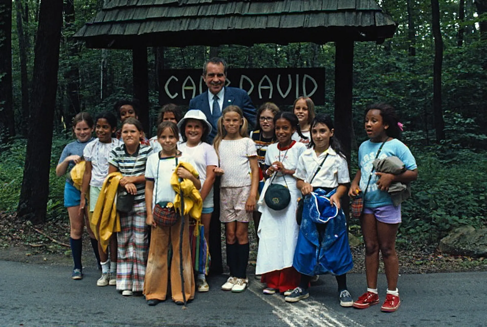 Girls of all races wearing colorful outfits pose with an older man wearing a suit in front of a sign reading 'Camp David.'