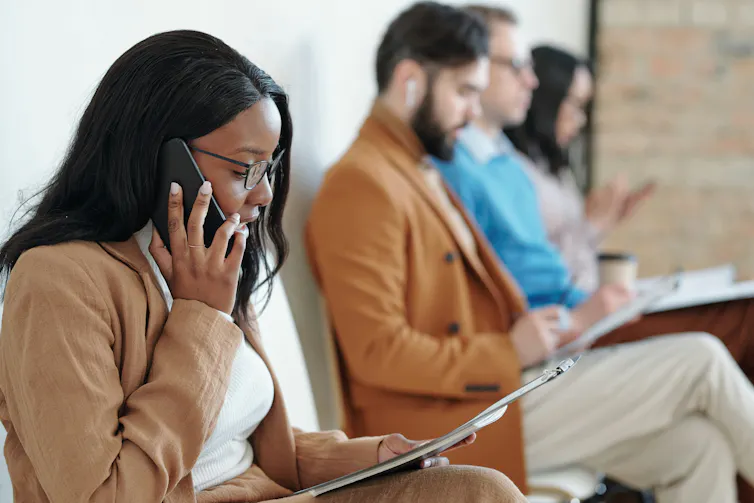 Four people sit on chairs holding clipboards, including a Black woman with long hair who talks on the phone.
