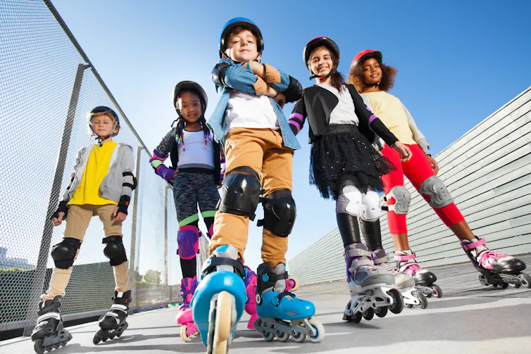 Five children on rollerblades wearing helmets and other safety gear pose in a skate park