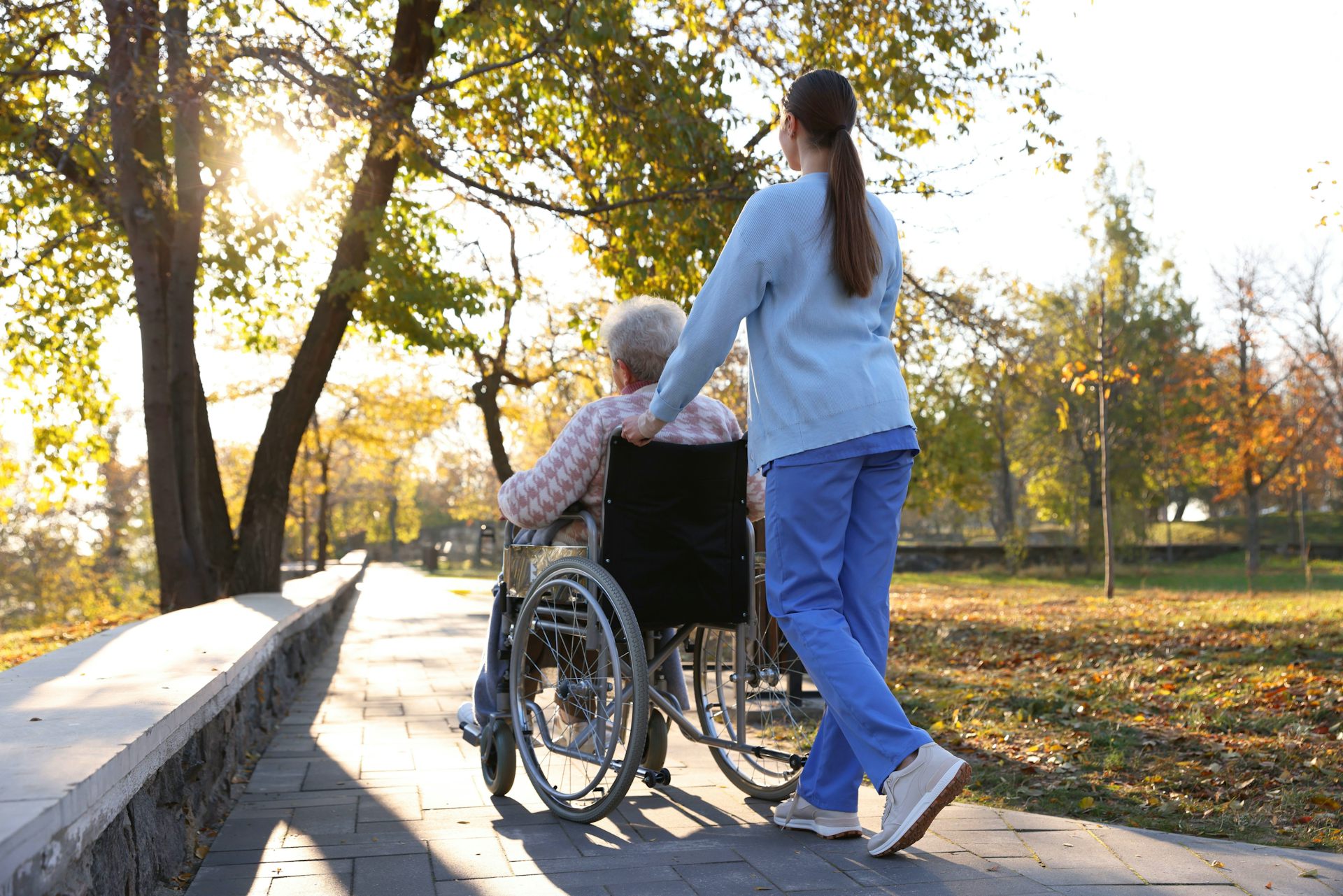 View from behind of a nurse pushing an elderly woman in a wheelchair, outdoors