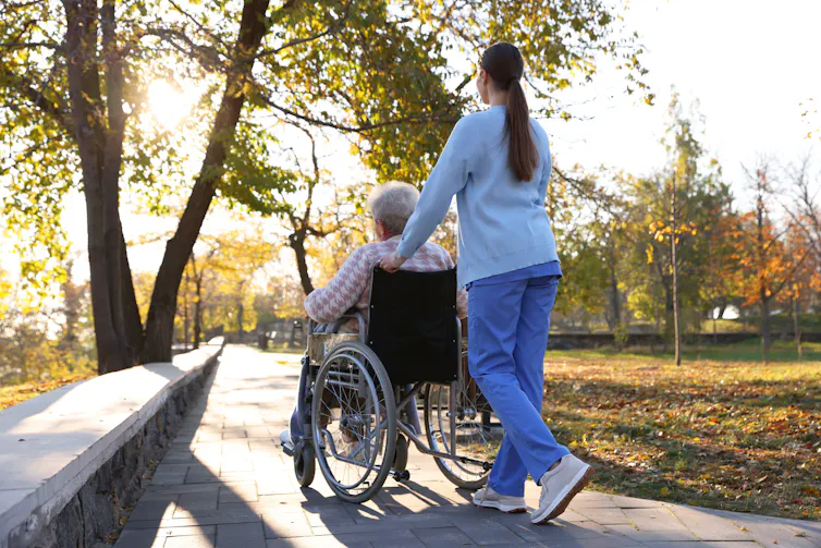 View from behind of a nurse pushing an elderly woman in a wheelchair, outdoors