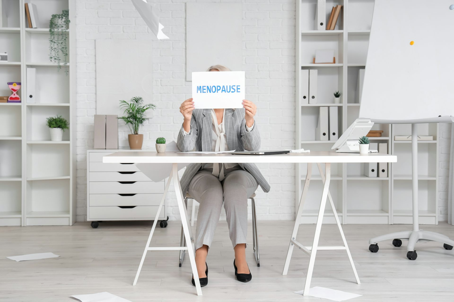 Femme assise à son bureau tenant devant son visage un panneau « Ménopause ».