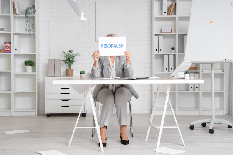 Femme assise à son bureau tenant devant son visage un panneau « Ménopause ».