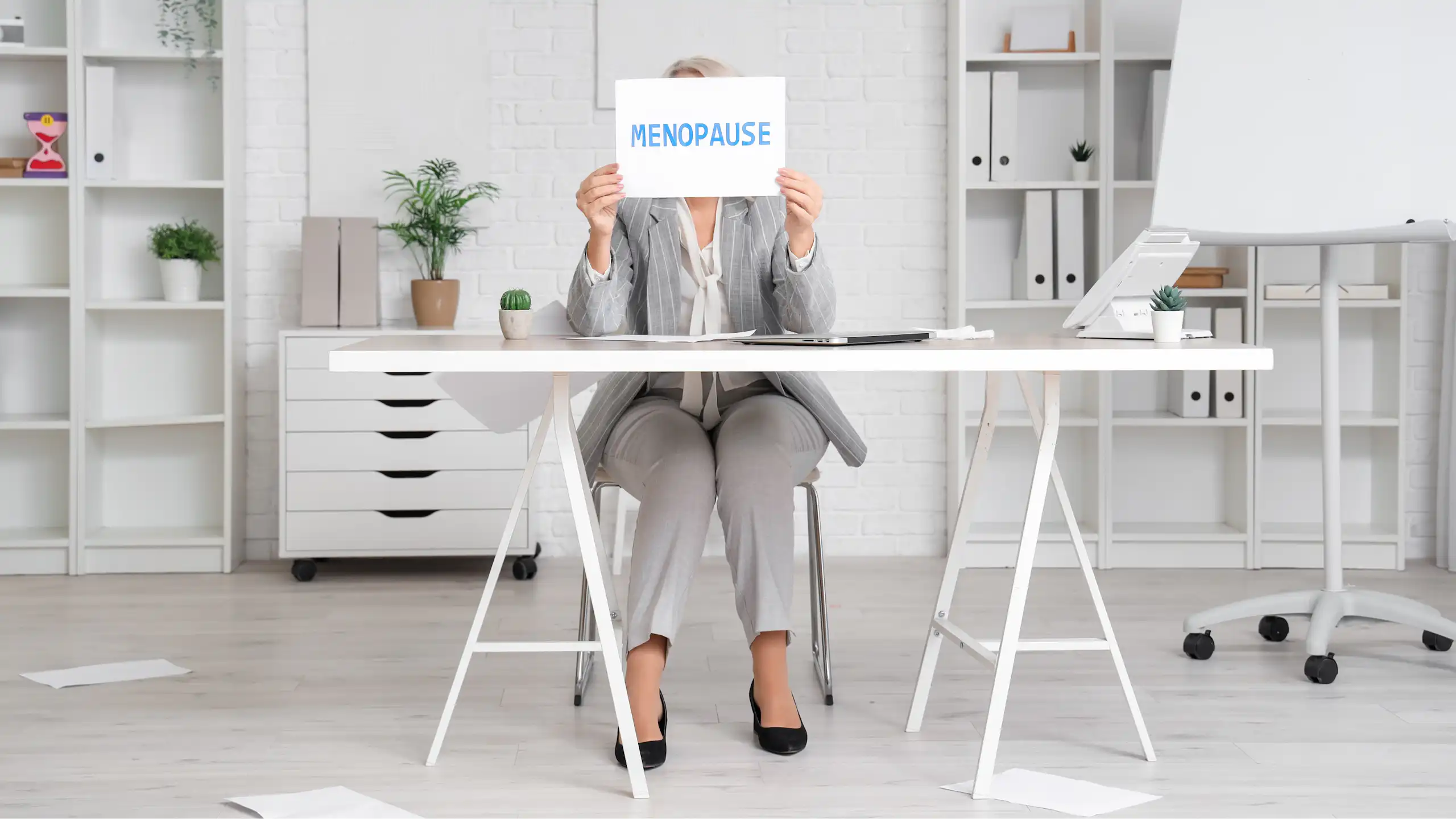 Femme assise à son bureau tenant devant son visage un panneau « Ménopause ».