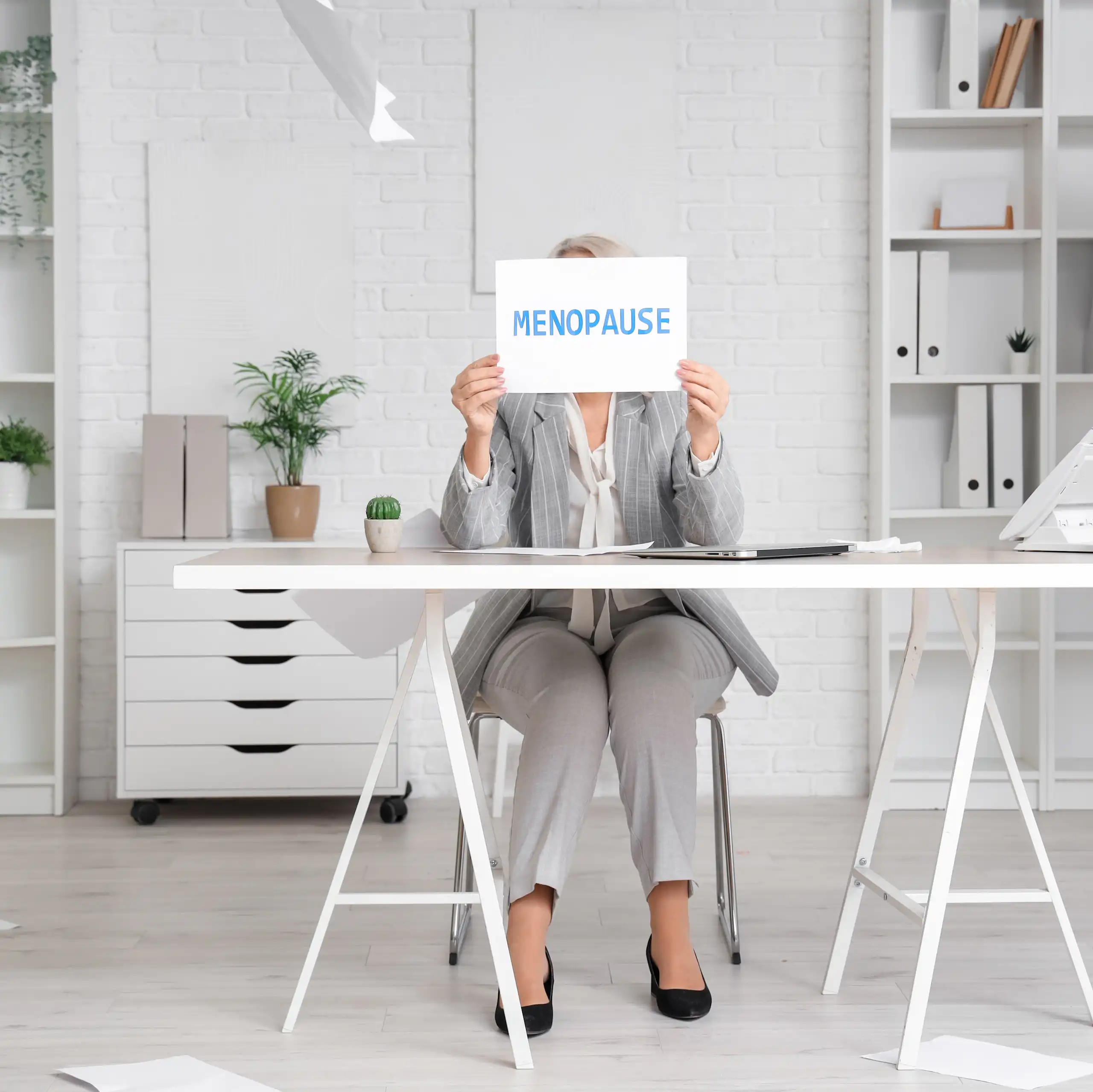 Femme assise à son bureau tenant devant son visage un panneau « Ménopause ».