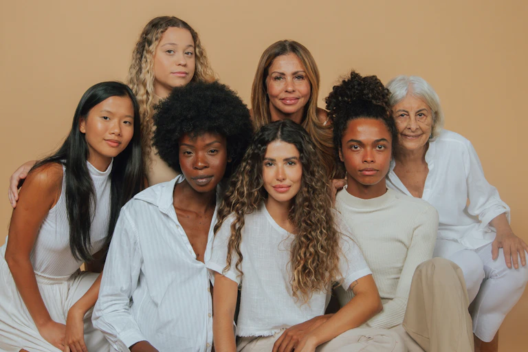 A group of women, who are very different in age, ethnicity and skin colour, stand together in a group and look on.