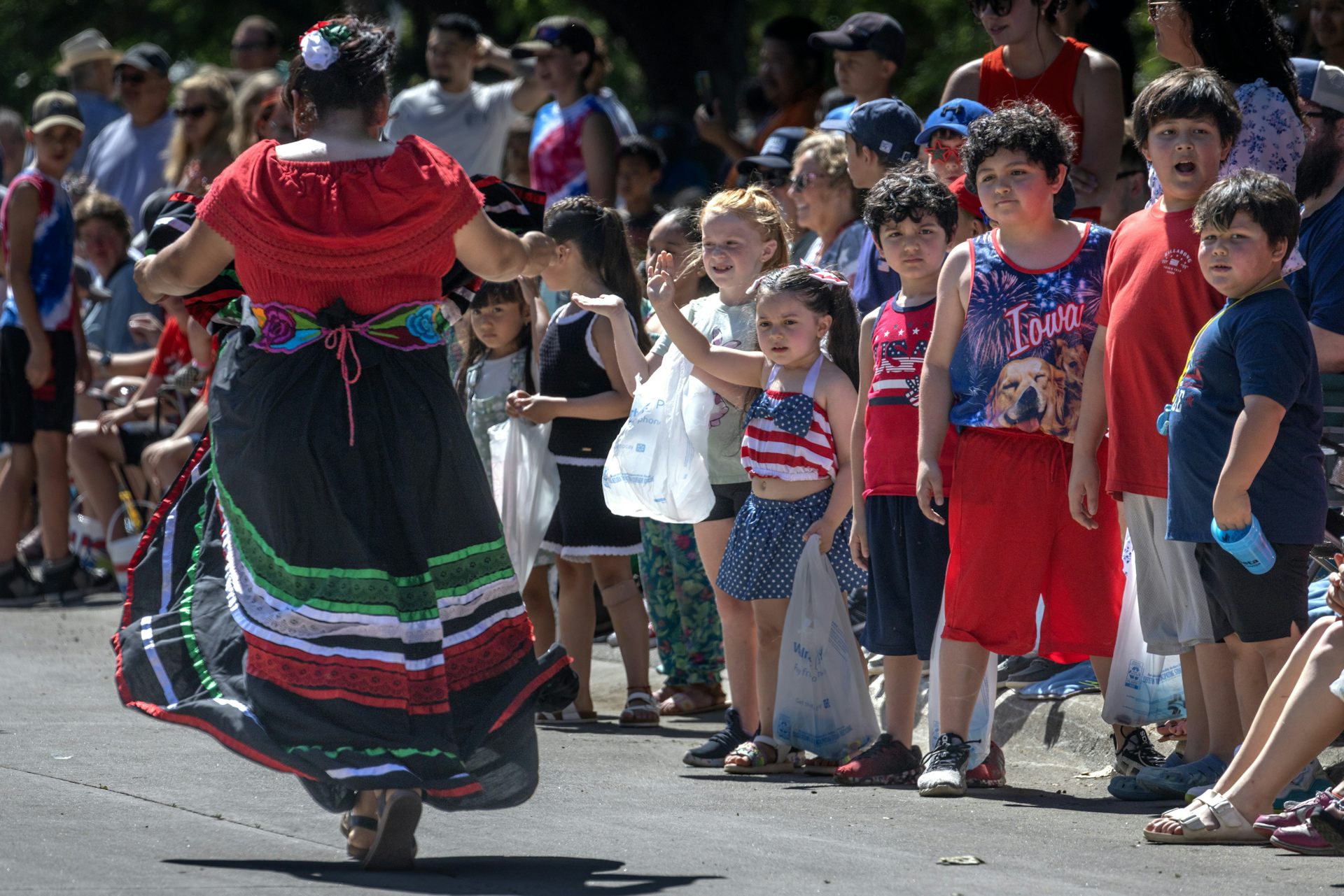 A person in a long red, black and green skirt walks past a row of children along a parade route.