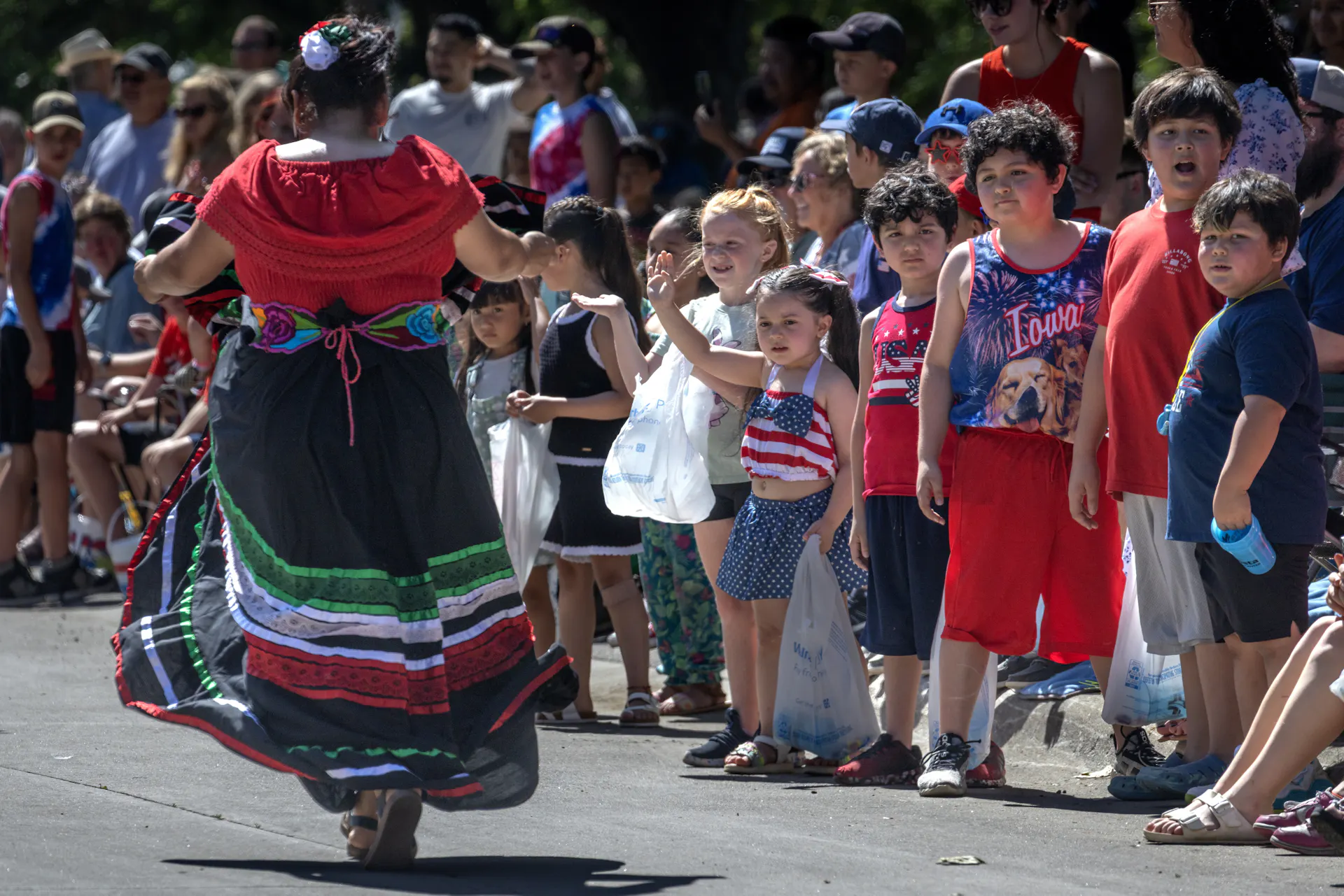 A person in a long red, black and green skirt walks past a row of children along a parade route.