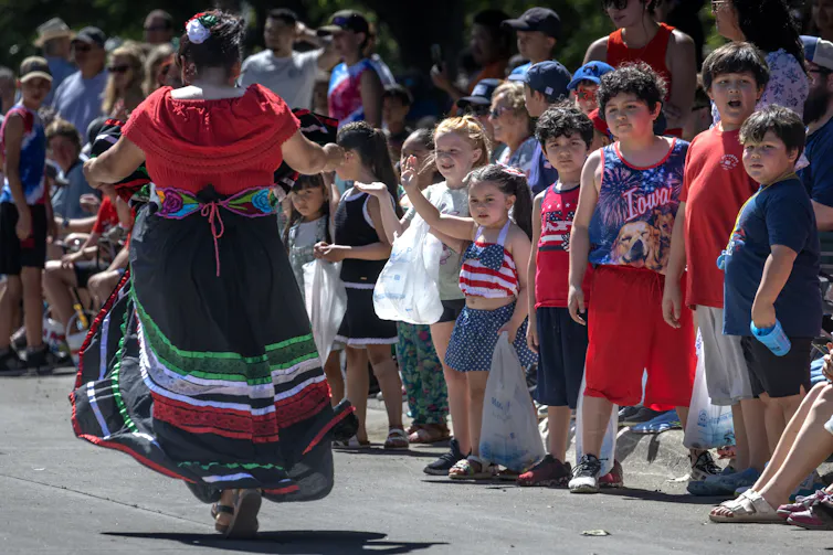 A person in a long red, black and green skirt walks past a row of children along a parade route.