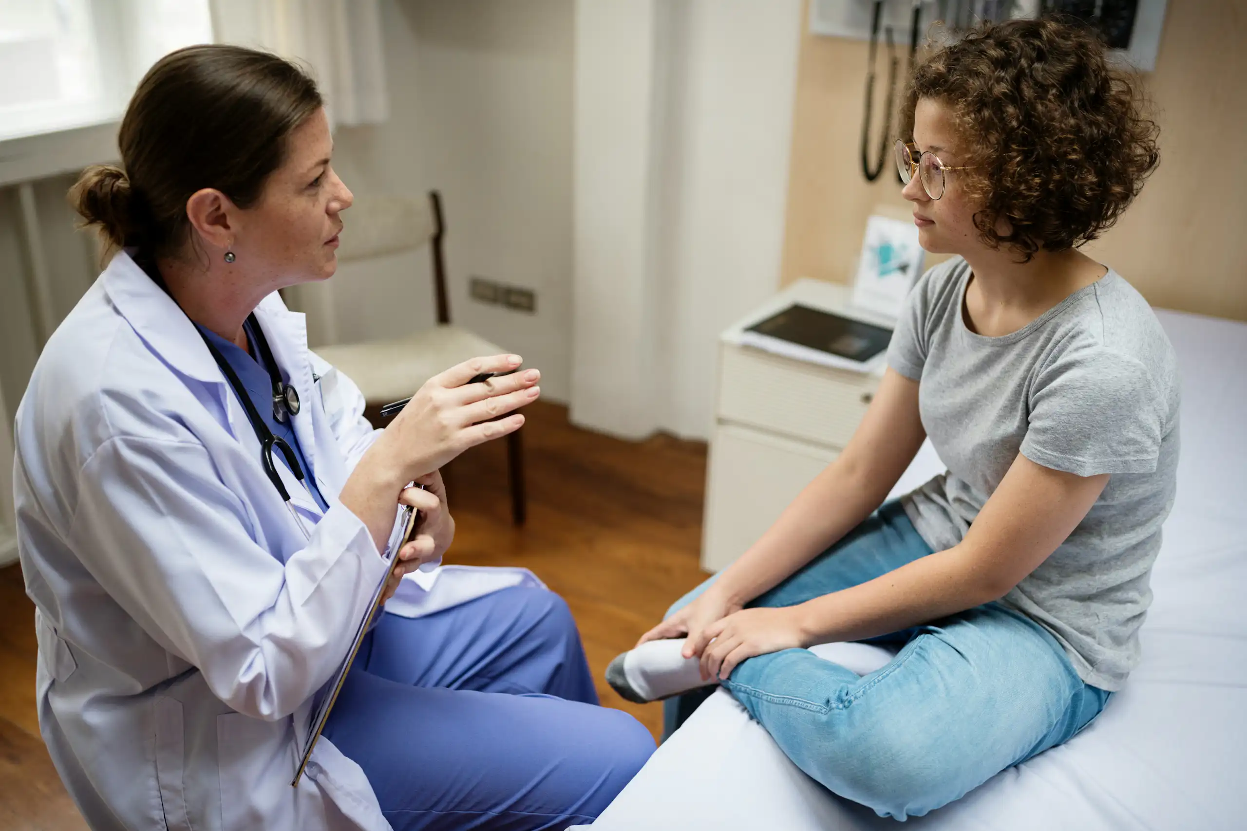 A woman health-care provider talking with a young woman in a grey T-shirt and jeans, sitting on an examination table.