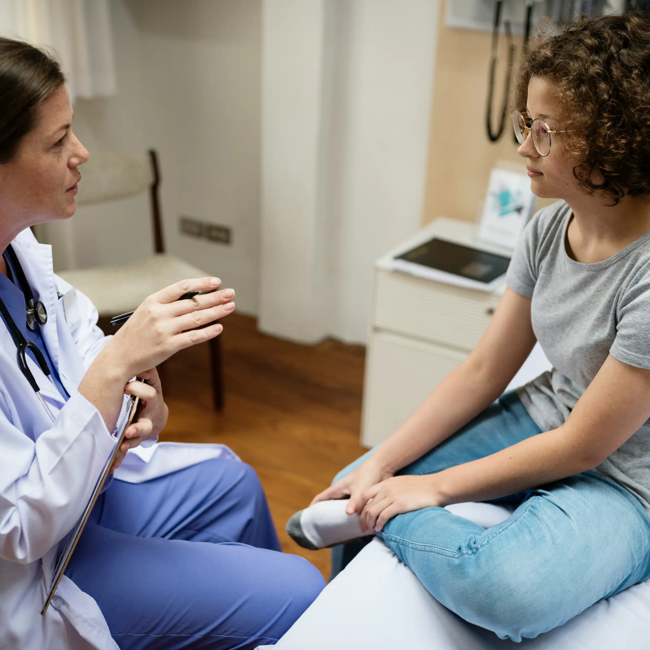 A woman health-care provider talking with a young woman in a grey T-shirt and jeans, sitting on an examination table.