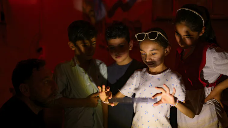 A group of children gather around a girl holding a strip of developed film in a darkroom. Photography