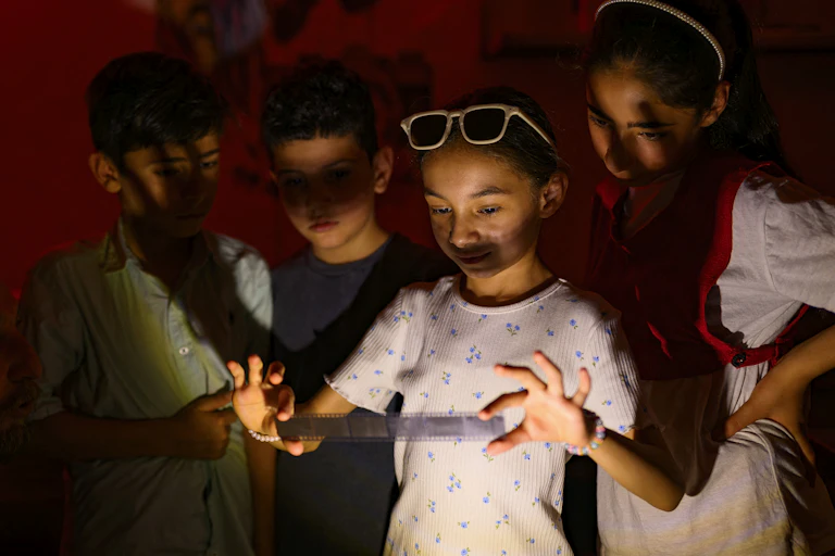 A group of children gather around a girl holding a strip of developed film in a darkroom.