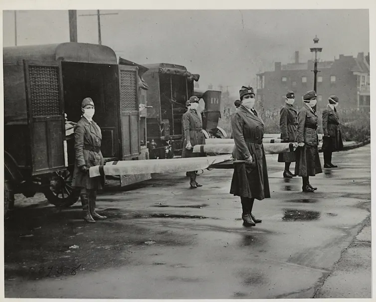 Mark-wearing nurses holding stretchers in front of military ambulances in a black and white photo from the 1918 flu pndemic