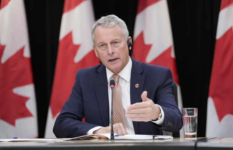 Un hombre blanco mayor con cabello blanco y traje oscuro habla detrás de un escritorio con una hilera de banderas canadienses detrás de él.