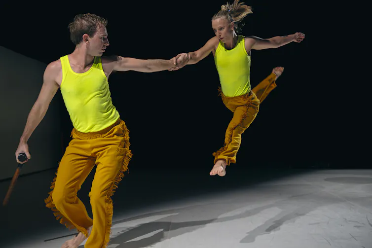 A woman jetés, holding a man's hand.