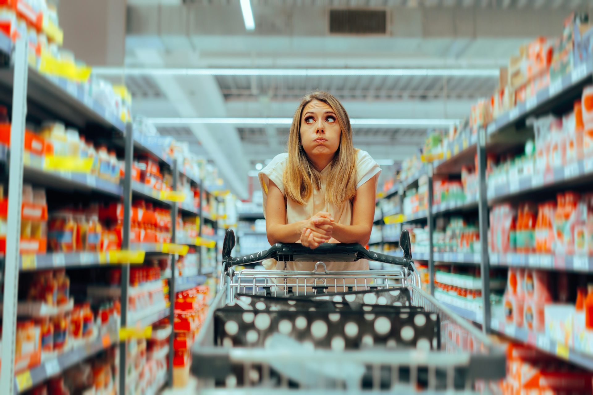 A woman looks confused while wheeling a shopping cart down a supermarket aisle.