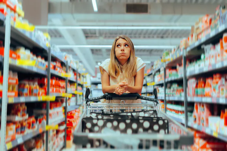 A woman looks confused while wheeling a shopping cart down a supermarket aisle.
